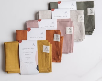 May include: Set of two linen napkins in mustard yellow, orange, pink, beige, and green. The napkins are folded and stacked on a white surface. The packaging includes the text "confetti mill" and "2 serviettes de table, Set of 2 napkins, 100% lin-linen".
