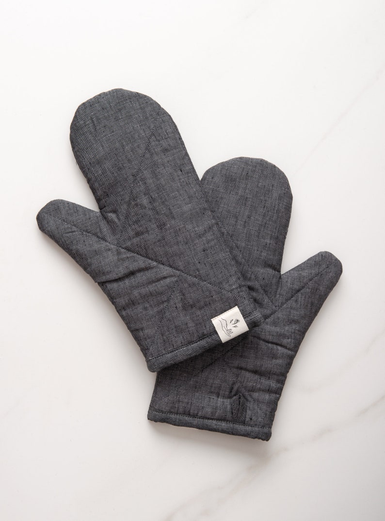 May include: A pair of gray oven mitts with a white label that says "The Good Hands" in black lettering.