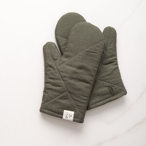 May include: A pair of olive green quilted oven mitts with a white label that says "La Isla".