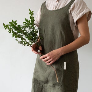 Può includere: Grembiule in lino verde oliva con un'ampia tasca frontale e spalline incrociate. Il grembiule è indossato sopra una camicia di colore chiaro. La persona tiene in mano un ramo di foglie. C'è una piccola etichetta sulla tasca.