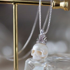 May include: A white pearl skull pendant necklace with a silver crown and clear cubic zirconia stones. The necklace is on a silver chain.
