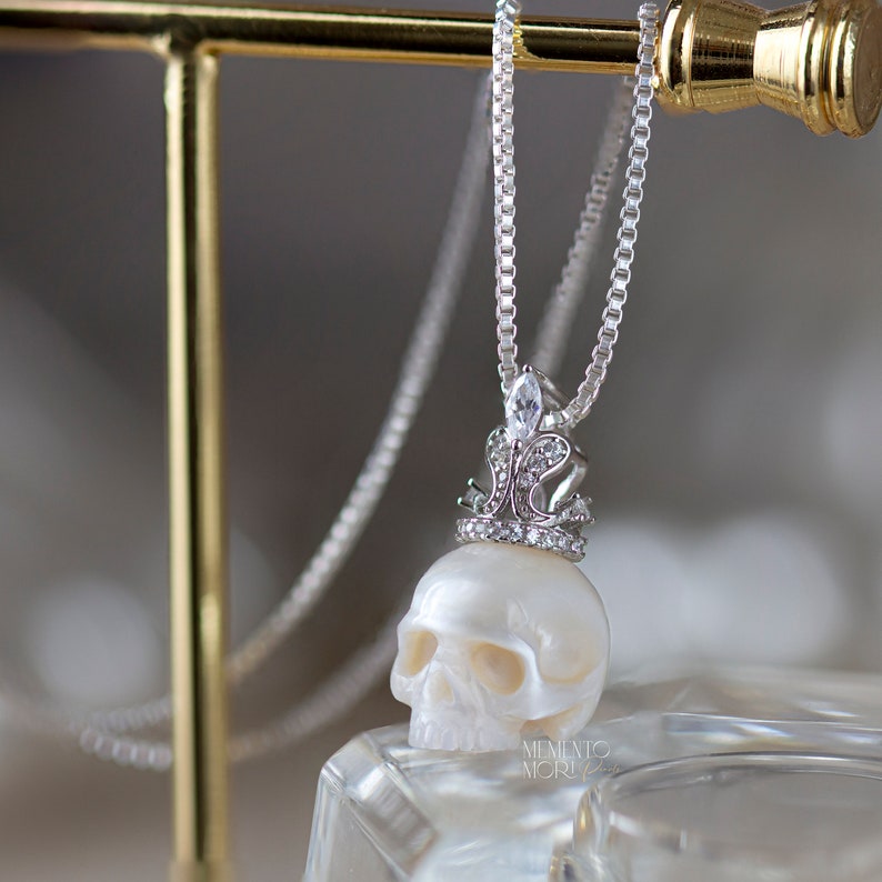 May include: A white pearl skull pendant necklace with a silver crown and clear cubic zirconia stones. The necklace is on a silver chain.