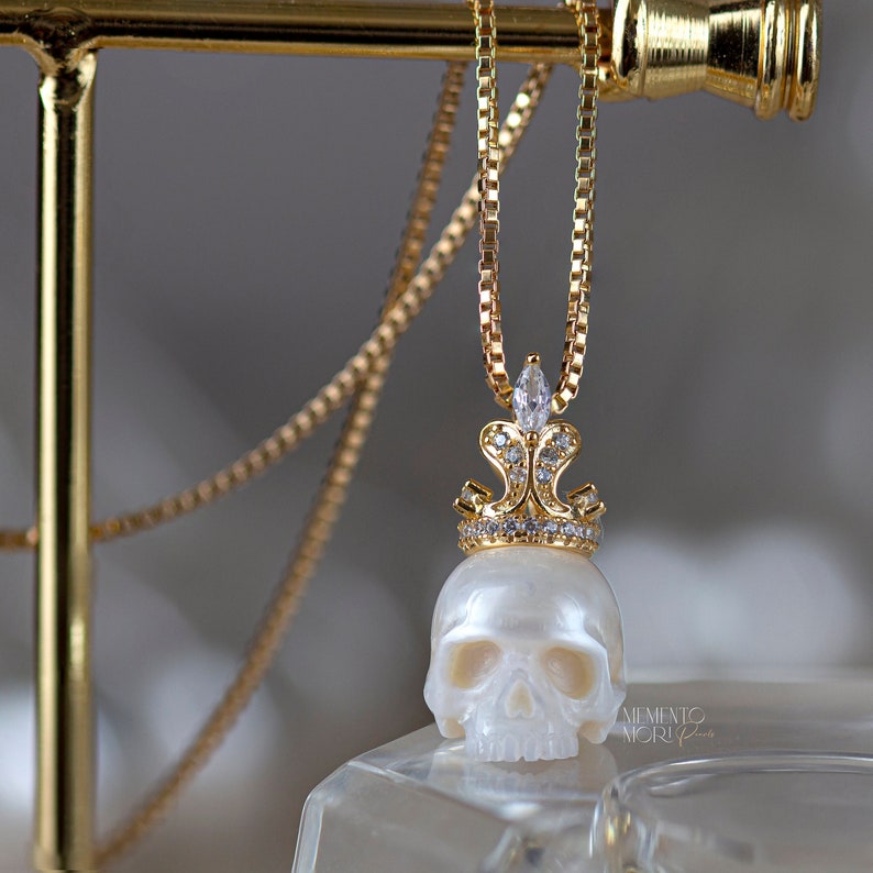 May include: A gold chain necklace with a white pearl skull pendant topped with a gold crown encrusted with clear cubic zirconia stones. The pendant is hanging from a gold chain on a gold display stand. The text "MEMENTO MORI" is visible in the bottom right corner of the image.