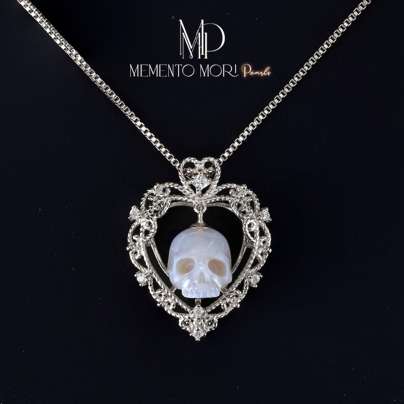 May include: A silver necklace with a heart-shaped pendant featuring a carved pearl skull. The pendant is adorned with small diamonds and the chain is a delicate box chain. The necklace is labeled "Memento Mori Pearl."