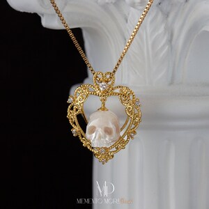 May include: A gold heart-shaped pendant necklace with a white skull carved from a pearl. The pendant is adorned with small diamonds and the text "MEMENTO MORI" is engraved on the bottom.