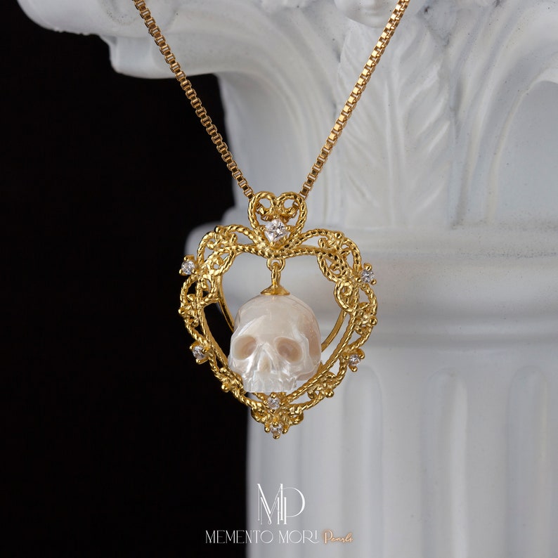 May include: A gold heart-shaped pendant necklace with a white skull carved from a pearl. The pendant is adorned with small diamonds and the text "MEMENTO MORI" is engraved on the bottom.