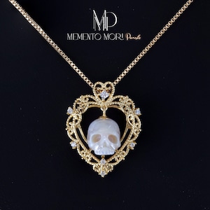 May include: A gold heart-shaped pendant with a white pearl skull inside. The pendant is on a gold chain and has small diamonds around the edge.