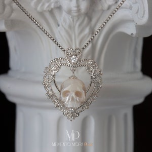 May include: A silver heart-shaped pendant necklace with a white pearl skull inside. The pendant is adorned with intricate details and small clear crystals. The necklace is on a silver chain. The text "MEMENTO MORI Pearl" is visible at the bottom of the image.