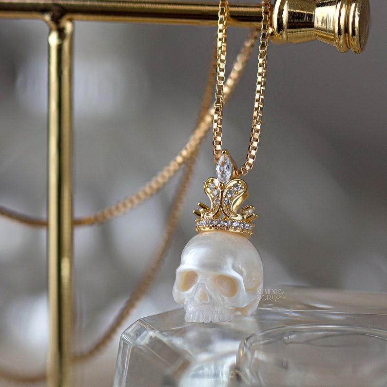 May include: A gold chain necklace with a white pearl skull pendant topped with a gold crown encrusted with clear rhinestones.