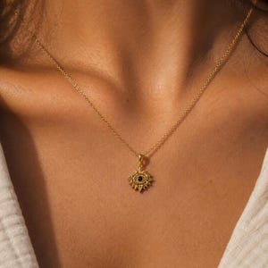 May include: Gold-colored necklace with a pendant featuring a sunburst design around a dark blue center. The necklace is worn around the neck, with the chain and pendant visible against the skin and a white shirt.