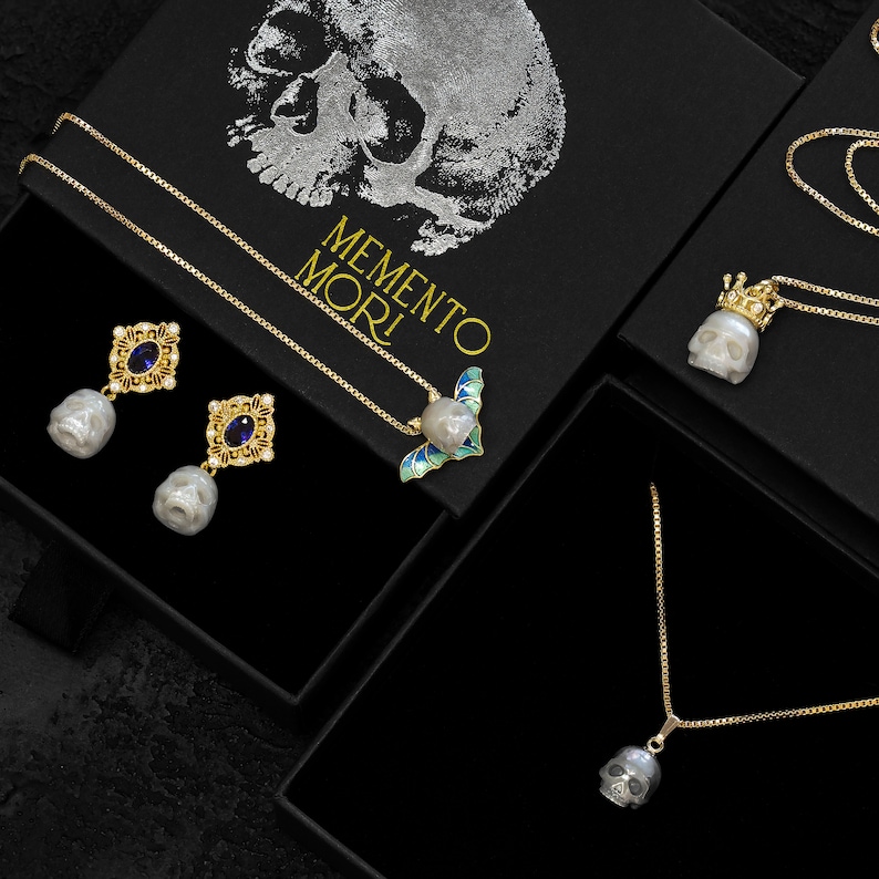 May include: Three gold necklaces with pearl skull pendants. One necklace features a crown, another a bat, and the third a plain skull. The necklaces are displayed in black boxes with the text "MEMENTO MORI".