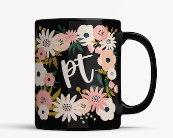 May include: Black ceramic mug with a floral wreath design in pink, white, and gold. The letters "pt" are in white script in the centre. The mug has a black handle and is suitable for hot or cold drinks.
