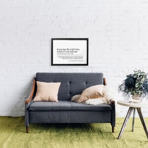 May include: A framed black and white print with the definition of "learning disabilities teacher consultant" on a white brick wall. A grey sofa with two cushions sits in front of the print. A small wooden table with a bouquet of grey and green flowers sits to the right of the sofa.