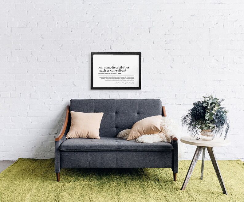 May include: A framed black and white print with the definition of "learning disabilities teacher consultant" on a white brick wall. A grey sofa with two cushions sits in front of the print. A small wooden table with a bouquet of grey and green flowers sits to the right of the sofa.