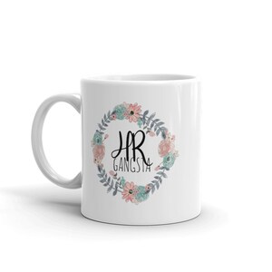 May include: White ceramic mug with a floral wreath design. The wreath is in shades of blue, pink, and green. The text "HR Gangsta" is in the center of the wreath.