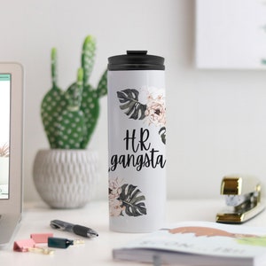 May include: White travel mug with a black lid, featuring the text "HR gangsta" in black script. The mug is decorated with floral and leaf designs in muted tones. A cactus and office supplies are in the background.