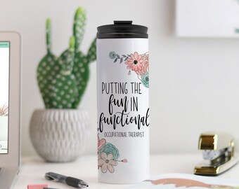 May include: White travel mug with a black lid, decorated with floral accents in pink, teal, and gray. The mug features the text "Putting the fun in functional Occupational Therapist."