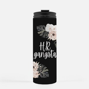 May include: Black travel mug with a black lid. The mug features the text "HR gangsta" in white script, along with floral and leaf accents in shades of white, pink, and green. The mug is designed to keep beverages hot or cold.