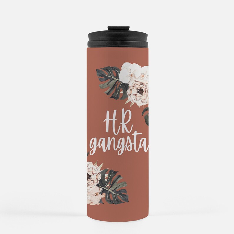May include: A terracotta-colored travel mug with a black lid. The mug features white script text that reads "HR gangsta" and is decorated with watercolor-style floral and leaf accents in white, green, and cream.