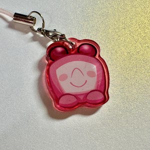 May include: A pink acrylic charm with a cartoon character design. The charm features a smiling face with a curved mouth and two small dots for eyes. It has a pink cord and a silver-colored clasp.