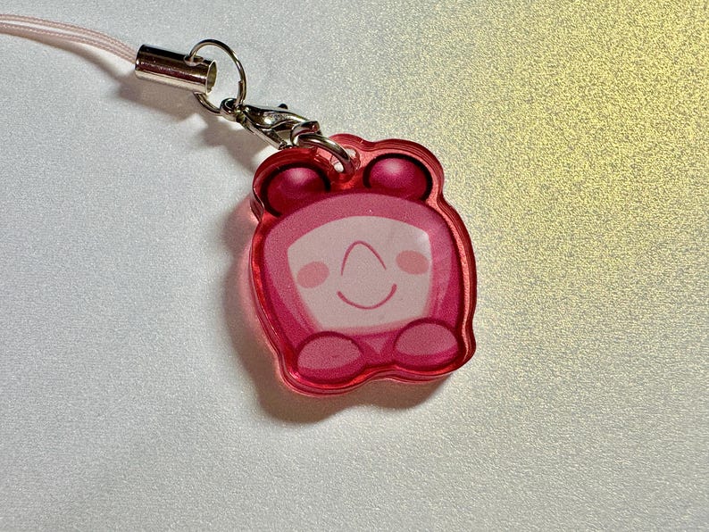 May include: A pink acrylic charm with a cartoon character design. The charm features a smiling face with a curved mouth and two small dots for eyes. It has a pink cord and a silver-colored clasp.