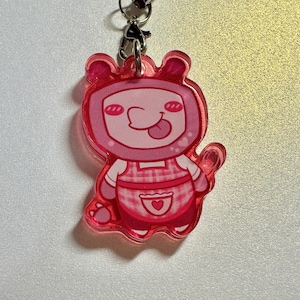 May include: A pink acrylic charm featuring a cartoon character with a square head, small ears, and a tongue sticking out. The character wears a pink and white checkered apron with a heart-shaped pocket. The charm is attached to a metal clasp and chain.