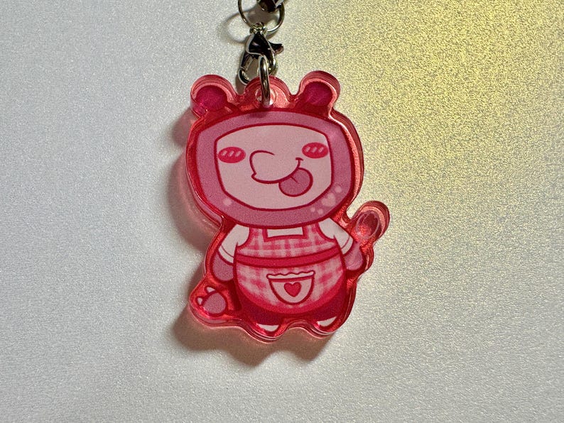 May include: A pink acrylic charm featuring a cartoon character with a square head, small ears, and a tongue sticking out. The character wears a pink and white checkered apron with a heart-shaped pocket. The charm is attached to a metal clasp and chain.