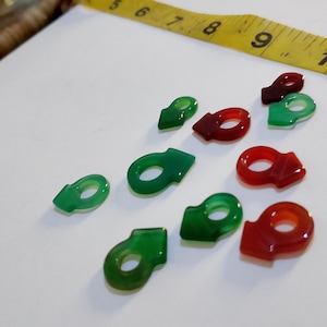 Puede incluir: Una colección de pequeños amuletos con forma de piedras preciosas en tonos verdes y rojos. Los amuletos están dispuestos sobre una superficie blanca con una cinta métrica amarilla en el fondo. Los amuletos tienen un diseño único de círculo abierto.