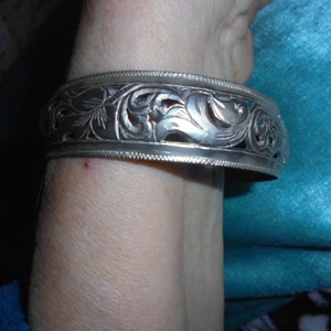 Puede incluir: Un brazalete de plata con un intrincado diseño floral. El brazalete se lleva en la muñeca.