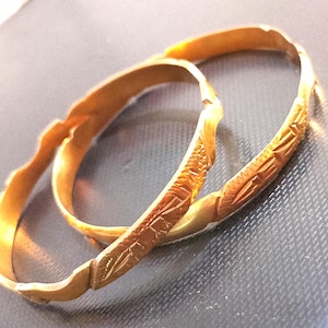 May include: Two gold-colored metal bangles featuring a textured, repeating leaf-like pattern engraved along the outer surface. The bracelets have a slightly wavy, irregular edge design and a polished finish that reflects light across the surface.