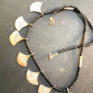 Könnte beinhalten: Eine Halskette mit einer Reihe kleiner schwarzer Perlen und einer goldenen zylindrischen Perle. An der Kette hängen sechs fächerförmige Metallanhänger, die jeweils mit einem geometrischen Muster aus Linien und Dreiecken graviert sind.