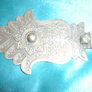 May include: A silver-colored metal ornament with intricate floral designs and a round, raised center. The ornament has a hook on the back.