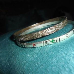 May include: Two silver bangles with intricate designs. The top bangle has a geometric pattern and the bottom bangle has a floral pattern with red and green enamel accents.