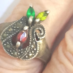 Puede incluir: Un anillo de estilo vintage con un diseño en forma de pera. El anillo presenta una piedra preciosa roja central, acentuada por piedras verdes y amarillas en forma de hoja. El anillo tiene un acabado plateado antiguo detallado.