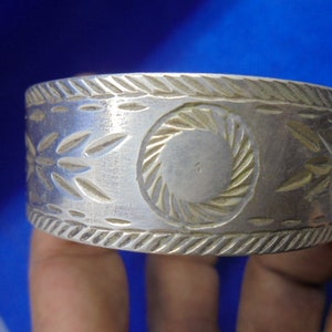 May include: A silver bracelet with an intricate design. The bracelet has a circular pattern in the center and a repeating leaf pattern on either side.