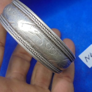 May include: A silver bracelet with an intricate design and a braided edge. The bracelet is engraved with a pattern of lines and swirls. The bracelet is labeled with the number "N45".