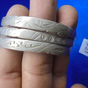 May include: Three silver bangles with intricate floral and swirl designs. The bangles are stacked on top of each other.