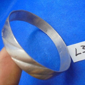 May include: A silver bangle bracelet with a twisted design. The bracelet is on a blue background with a white tag that says "L37".