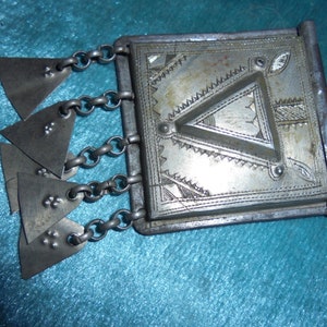 May include: A silver metal charm with a geometric design and a chain with triangular pendants. The charm has a hinged lid and a small loop on the side.
