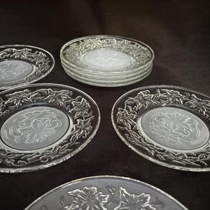 May include: Clear glass oval plates with a floral design around the rim and a central embossed floral pattern. Several plates are stacked, while others are arranged individually, showcasing the design.