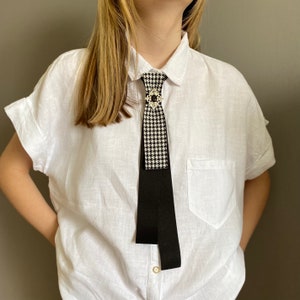 May include: A black and white houndstooth tie with a black ribbon, secured by a decorative brooch with a black stone. The tie is worn over a white button-down shirt.