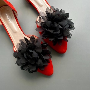 May include: A pair of red pointed-toe flats featuring a large black sheer fabric flower on the toe of each shoe. The interior of the shoes is a light beige color with the brand name Gino Rossi printed in white on the heel area of the insole.