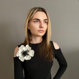 May include: A large white and black flower brooch pinned to a black long-sleeve top. The flower has multiple white petals surrounding a black center. The top has cold shoulder cutouts.