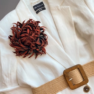 Puede incluir: Un blazer blanco con un cinturón tejido marrón y una hebilla cuadrada de madera grande. Un broche de flores rojo oscuro está prendido al blazer. El blazer tiene una etiqueta CA Premium.