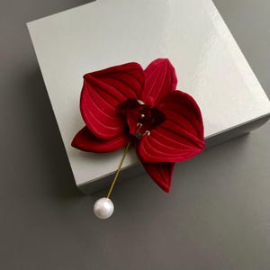 May include: A red velvet orchid brooch with a pearl accent. The brooch is attached to a gold pin.