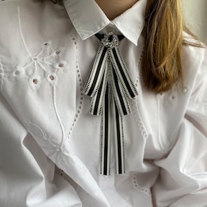 Könnte beinhalten: Eine schwarz-weiß gestreifte Schleife mit dekorativem Strass- und Perlenverschluss. Die Fliege ist an einem weißen Hemd mit Kragen und Lochstickerei befestigt.