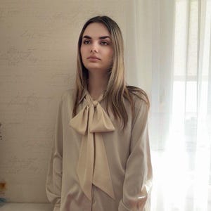 May include: A beige long-sleeve blouse with a large bow tie. The blouse has a classic collar and buttoned cuffs. The background is a light-colored wall with faint script writing.