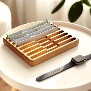 May include: A wooden watch band organizer with multiple slots, holding several pastel-colored watch bands. A black smartwatch with a black band rests on the table. The organizer is made of light-colored wood.