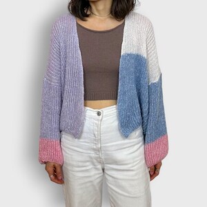 Puede incluir: Un cárdigan de bloques de color en tonos lavanda, blanco y azul, con puños rosas. El suéter abierto está hecho de un material de punto grueso. La modelo lleva un top corto marrón y pantalones blancos.