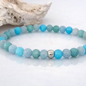 Könnte beinhalten: Ein Perlenarmband mit einem sich wiederholenden Muster aus glatten, runden Edelsteinperlen in verschiedenen Schattierungen von Hellblau, Türkis und Blaugrün. Eine einzelne, polierte, reflektierende silberfarbene Metallperle ist mittig platziert.